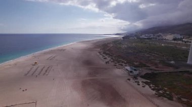 Fuerteventura 'da bulutlu bir gökyüzü altında Morro Jable' ı yakalayan muhteşem hava görüntüleri. Resim, seyahat, doğa ve macera fotoğrafçılığı için mükemmel olan altın kumlar, modern mimari ve yuvarlanan tepeler arasındaki zıtlığı sergiliyor.. 