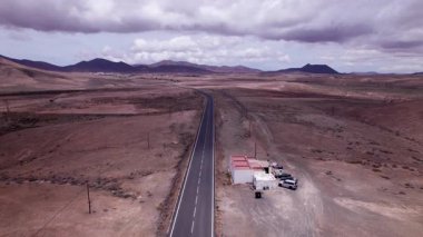 Fuerteventura 'nın uçsuz bucaksız, çorak tepelerini keserek düz bir yol yakalayan muhteşem hava görüntüleri. Görüntü, kırmızı toprağın, dağınık bitki örtüsünün ve uzak dağların, seyahat ve doğa fotoğrafçılığı için mükemmel güzelliklerini sergiliyor.. 