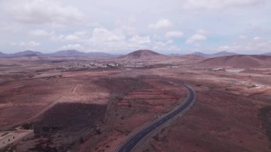Fuerteventura 'daki dramatik kırmızı volkanik manzarayı kesen asfaltlı yolun geniş manzarası. Sahne, çorak güzelliği, derinliği ve seyahat potansiyelini yakalar..