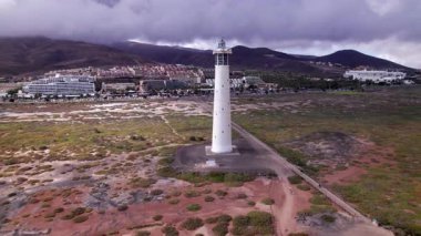 Dinamik hava görüntüleri Fuerteventura 'daki Morro Jable' ın görkemli dağlarının yakınındaki ikonik deniz fenerini gösteriyor. Görüntü, kızıl toprağın, dağınık bitki örtüsünün ve uzak tepelerin saf güzelliğini yakalıyor. Seyahat için ideal, doğa fotoğrafçılığı.. 