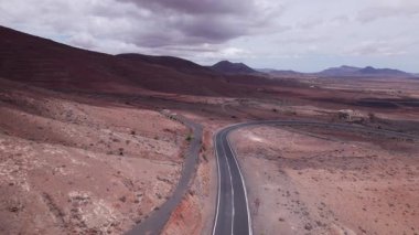 Fuerteventura 'daki dramatik kırmızı volkanik manzarayı kesen asfaltlı yolun geniş manzarası. Sahne, çorak güzelliği, derinliği ve seyahat potansiyelini yakalar..
