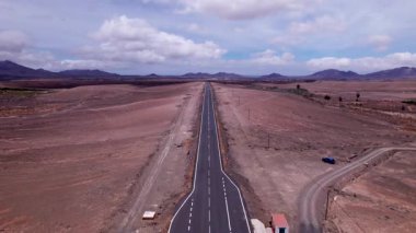 Dinamik hava çekimi Fuerteventura 'daki görkemli uzak dağlara doğru uzanan düz bir yol gösteriyor. Görüntü, yapılandırılmış yol ile engebeli arazi arasındaki kontrastı, seyahat ve yaşam tarzı fotoğrafçılığı için ideal olanı yakalıyor.. 