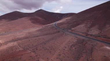 Fuerteventura 'daki dramatik kırmızı volkanik manzarayı kesen asfaltlı yolun geniş manzarası. Sahne, çorak güzelliği, derinliği ve seyahat potansiyelini yakalar..