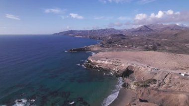 Dinamik hava çekimi Fuerteventura 'daki La Pared yakınlarında turkuaz sulara bakan dramatik uçurumları gösteriyor. Görüntü, kırmızı toprağın, dağınık bitki örtüsünün ve seyahat fotoğrafçılığı için ideal uzak tepelerin saf güzelliğini yakalar.. 