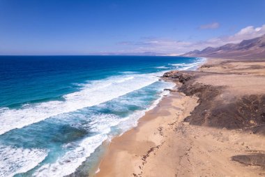 Dinamik hava çekimi Fuerteventura 'daki Playa de Coffee' de Roque del Moro yakınlarındaki güzel plajı ve kayalık sahili gösteriyor. Görüntü, altın kumlar, turkuaz sular ve seyahat fotoğrafçılığı için ideal engebeli arazi arasındaki zıtlığı yakalıyor. 