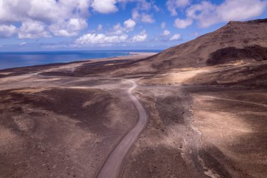 Jandia, Fuerteventura 'da dramatik bir gökyüzü altında düz bir yol yakalayan muhteşem hava görüntüleri. Görüntü, kırmızı toprağın, dağınık bitki örtüsünün ve uzak tepelerin katıksız güzelliğini sergiliyor. Bu da onu seyahat ve macera fotoğrafçılığı için ideal kılıyor.. 