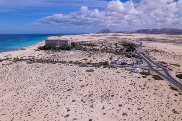 Fuerteventura 'daki Corralejo' nun engin kumlu arazisine yerleştirilmiş modern bir otelin detaylı hava görüntüleri. Görüntü, kırmızı toprağın, dağınık bitki örtüsünün ve uzak tepelerin katıksız güzelliğini vurgular.. 