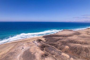 Dinamik hava görüntüleri, Fuerteventura 'daki Playa de Cofete' de Roque del Moro yakınlarındaki nefes kesici plajı ve uçurumları gösteriyor. Görüntü, altın kumlar, turkuaz sular ve seyahat fotoğrafçılığı için ideal engebeli arazi arasındaki zıtlığı yakalıyor.. 