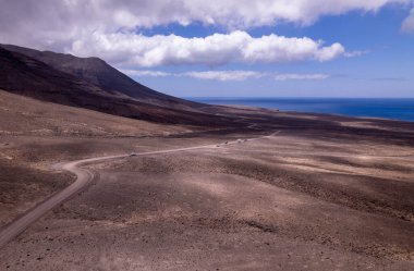Fuerteventura 'daki Jandia' nın uçsuz bucaksız, çorak arazisini kesen dolambaçlı bir yolu yakalayan muhteşem hava görüntüleri. Görüntü, kırmızı toprağın, dağınık bitki örtüsünün ve uzak dağların katıksız güzelliğini gösteriyor.. 