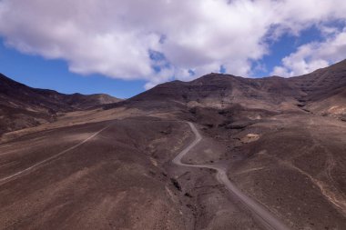 Detaylı hava görüntüleri Fuerteventura 'da Jandia' nın açık alanlarını kesen dolambaçlı bir yolu gösteriyor. Görüntü, kırmızı toprağın, dağınık bitki örtüsünün ve seyahat ve macera içeriği için mükemmel olan uzak dağların saf güzelliğini vurgular..