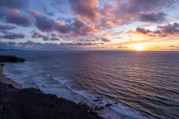 La Pared, Fuerteventura yakınlarındaki volkanik bir kıyı şeridinde canlı bulutlar ve dalgalarla altın gün batımı. 