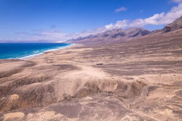 Dinamik hava çekimi Fuerteventura 'daki Playa de Cofete' de Roque del Moro yakınlarındaki turkuaz sulara bakan dramatik uçurumları gösteriyor. Görüntü, kırmızı toprağın, dağınık bitki örtüsünün ve uzak tepelerin saf güzelliğini yakalar..