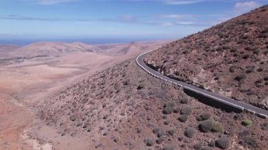 Detaylı hava görüntüleri, Fuerteventura 'nın uçsuz bucaksız, çorak arazisini kesen dolambaçlı bir yolu yakalıyor. Görüntü, kırmızı toprağın, dağınık bitki örtüsünün ve uzak tepelerin katıksız güzelliğini vurguluyor. Bu da onu seyahat içeriği için ideal kılıyor..