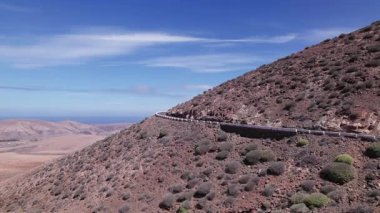 Detaylı hava görüntüleri, Fuerteventura 'nın uçsuz bucaksız, çorak arazisini kesen dolambaçlı bir yolu yakalıyor. Görüntü, kırmızı toprağın, dağınık bitki örtüsünün ve uzak tepelerin katıksız güzelliğini vurguluyor. Bu da onu seyahat içeriği için ideal kılıyor..