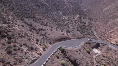 Dinamik hava çekimi Fuerteventura 'daki yuvarlanan tepelerin yakınında virajlı bir yol gösteriyor. Görüntü, yapılandırılmış yol ile engebeli arazi arasındaki zıtlığı, seyahat, emlak ve yaşam tarzı fotoğrafçılığı için ideal olanı yakalıyor.. 