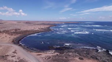 Fuerteventura 'da ufka doğru uzanan bir kıyı şeridi görüntüsü. Görüntü, kırmızı toprağın, dağınık bitki örtüsünün ve uzak tepelerin, seyahat, doğa ve macera fotoğrafçılığı için mükemmel güzelliklerini sergiliyor..