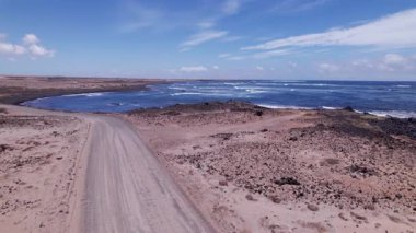 Dinamik hava çekimi Fuerteventura 'daki turkuaz suların yakınında dolambaçlı bir yol gösteriyor. Görüntü, yapılandırılmış yol ile engebeli arazi arasındaki zıtlığı, seyahat, emlak ve yaşam tarzı fotoğrafçılığı için ideal olanı yakalıyor.. 