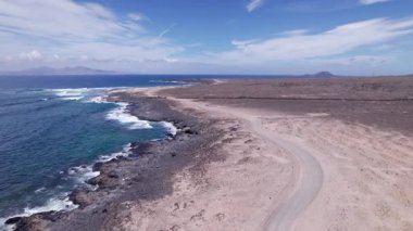 Fuerteventura 'da okyanus kenarında dönen bir kıyı yolunu yakalayan muhteşem hava görüntüleri. Görüntü, kırmızı toprağın, dağınık bitki örtüsünün ve uzak tepelerin, seyahat, doğa ve macera fotoğrafçılığı için mükemmel güzelliklerini sergiliyor.. 