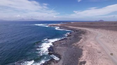 Fuerteventura kıyısındaki engin kurak araziyi gösteren detaylı hava görüntüleri. Görüntü, kırmızı toprağın, dağınık bitki örtüsünün ve uzak tepelerin katıksız güzelliğini vurguluyor. Bu da onu seyahat, doğa ve macera içeriği için ideal kılıyor.. 