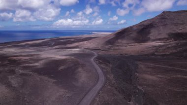 Fuerteventura 'da ufka doğru uzanan bir kıyı şeridi görüntüsü. Görüntü, kırmızı toprağın, dağınık bitki örtüsünün ve uzak tepelerin, seyahat, doğa ve macera fotoğrafçılığı için mükemmel güzelliklerini sergiliyor..