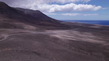 Uçsuz bucaksız Fuerteventura 'nın kurak arazisini kesen dolambaçlı bir yolu yakalayan muhteşem hava görüntüleri. Görüntü, kırmızı toprağın, dağınık bitki örtüsünün ve uzak dağların, seyahat ve doğa fotoğrafçılığı için mükemmel güzelliklerini sergiliyor..