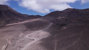 Dinamik hava görüntüleri Fuerteventura 'daki görkemli dağların yakınında bir kıyı yolu gösteriyor. Görüntü, turkuaz suların doğal güzelliği ile yapılandırılmış yol arasındaki zıtlığı yakalıyor. Seyahat, emlak ve yaşam tarzı fotoğrafçılığı için ideal.