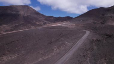Dinamik hava çekimi Fuerteventura 'daki yuvarlanan tepelerin yakınında virajlı bir yol gösteriyor. Görüntü, yapılandırılmış yol ile engebeli arazi arasındaki zıtlığı, seyahat, emlak ve yaşam tarzı fotoğrafçılığı için ideal olanı yakalıyor.. 
