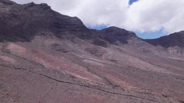 Kahveye, Fuerteventura 'daki dağların yakınındaki kurak araziyi gösteren muhteşem hava görüntüleri. Görüntü kırmızı toprağın, dağınık bitki örtüsünün ve uzak tepelerin katıksız güzelliğini sergiliyor. Yolculuk ve macera fotoğrafçılığı için mükemmel..