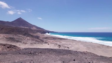 Fuerteventura 'daki Coffee' deki kayalık kayalıkların yakınındaki çarpıcı sahili dramatik bir şekilde görüntülüyor. Görüntü, kırmızı toprağın, dağınık bitki örtüsünün ve uzak tepelerin katıksız güzelliğini vurguluyor. Bu da onu seyahat, doğa ve macera içeriği için ideal kılıyor..