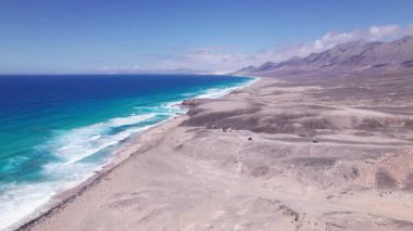 Dinamik hava görüntüleri Fuerteventura 'daki Coffee' deki kumsalın yakınındaki kurak araziyi yakalıyor. Görüntü, kırmızı toprağın, dağınık bitki örtüsünün ve uzak tepelerin katıksız güzelliğini vurgular. Bu da onu seyahat için ideal kılar, doğa içeriği..