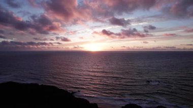 Sakin okyanus sularının yakınında çarpıcı günbatımını gösteren dramatik bir hava çekimi. Görüntü gökyüzünün canlı renklerini yansıtır, yansıtıcı suyu ve uzak tepeleri, seyahat, emlak ve yaşam tarzı fotoğrafçılığı için idealdir.