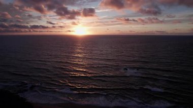 Turkuaz sularda nefes kesen gün batımını gösteren dramatik hava çekimi. Görüntü, gökyüzünün canlı renklerini, sakin okyanusları ve uzak tepeleri, seyahat, gayrimenkul ve yaşam tarzı fotoğrafçılığı için idealleri yakalıyor.