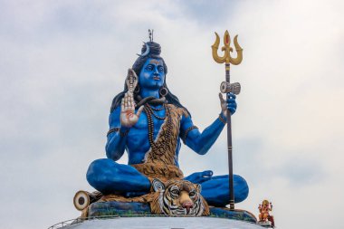 Lord Shiva 'nın mavi heykeli kaplan derisinden yapılmış, mızrak tutuyor ve kutsal bir jest sergiliyor. Yılanlar ve boncuklarla süslenmiş bu canlı heykel, berrak bir gökyüzüne karşı görkemli bir şekilde duruyor.