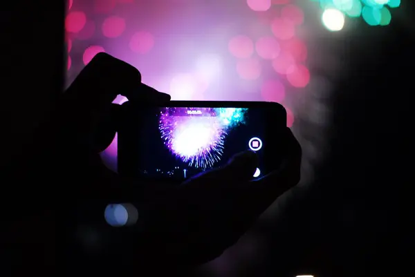 Bir insan cep telefonunu gökyüzünü aydınlatan renkli havai fişeklerin fotoğrafını çekmek için kullanıyor.