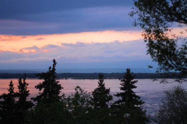 Uzun çam ağaçlarıyla çerçevelenmiş bir nehrin üzerinde sakin bir günbatımı manzarası ihtişamlı doğasını gözler önüne seriyor.