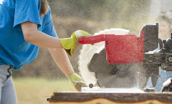 Tanımlanamayan, parlak mavi tişörtlü, minyon bir kadın gündüz inşaat alanında büyük, kırmızı bir testere kullanarak bir tahta parçasını kesiyor..