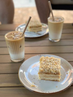 Tabaktaki bir dilim pasta buzlu kahve bardaklarıyla eşleştirilir, ahşap bir masaya kurulur, sıcak ve davetkar bir kafe deneyimini anımsatır.