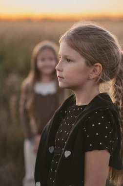 Uzun saçlı genç bir kız gün batımında bir tarlada duruyor, siyah bir benekli tişört giyiyor, arka planda hafifçe bulanık bir başka kız, sakin bir atmosferi çağrıştırıyor.