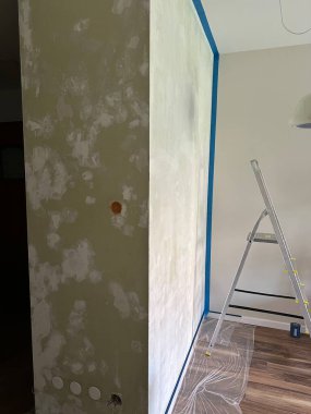 Duvar yenileniyor, tamamlanmamış yüzey ve boyacı merdiveni yer alıyor, ev iyileştirme projesini ve boyama hazırlıklarını vurguluyor