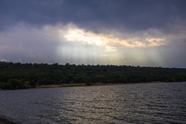 Güneşin ışınları nehirden gelen suya yansıyor. Yağmur bulutlarının arkasından çıkıyor..
