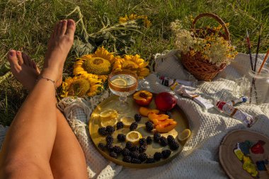 Bilezik takmış, üzerinde meyve, tarla çiçekleri ve boyamak için nesneler olan bir battaniyenin üzerinde oturan rahat bir bacak görüntüsü..