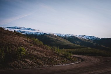 Şafak vakti karla kaplı Alaska dağlarına giden eğimli bir yolun huzur veren güzelliğini keşfedin. Macera, seyahat ve doğa temaları için mükemmel. Alaska 'nın görkemli vahşi doğasını gözler önüne seriyor..