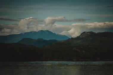 Alaska 'nın engebeli manzarasının hayranlık uyandıran güzelliğini bu çarpıcı görüntüyle yakalayın. Görkemli dağlar, yemyeşil ormanlar ve dramatik bir gökyüzü altında sakin sular..