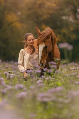 Bir kadın ve Lusitano atı sonbaharda bir kır çiçeğinin çayırında sarılıp kaynaşıyorlar.