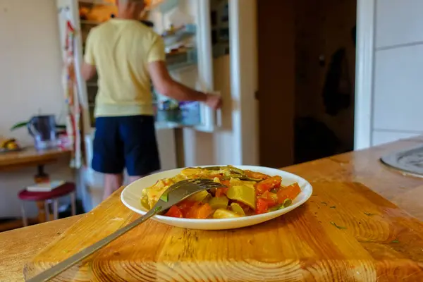 A plate of food on the table. In the background, a man opened the refrigerator