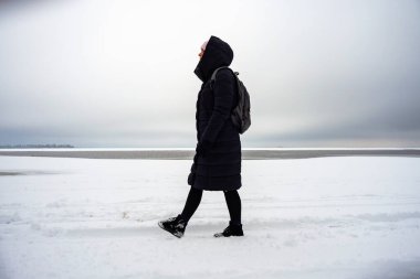 Karlı bir nehir kıyısında bir kadın yürüyor.