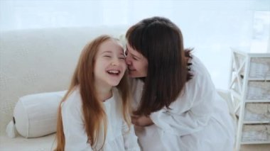 Genç anne ve güzel kızıl saçlı 6 yaşındaki kız sabah evde eğleniyorlar. Anne gülerken mutlu kızını öpüyor, 4k video.