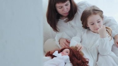 Anne ve küçük kızı kanepede oturuyor ve kız bir bebekle oynuyor. Çocuk kız, oyuncak bebeğin dolusunu tarar ve kızına bakarken kadın gülümser. Duvarın arkasından çevirme, 4K video