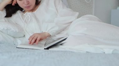 Genç esmer bir kadının portresi, yatakta yatarken kitap okuyor, 4K görüntü.