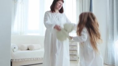 Genç anne ve küçük tatlı kız eski moda beyaz pijama giyip evde eğleniyorlar. Kızı olan bir kadın oturma odasında el ele tutuşuyor, 4K video.